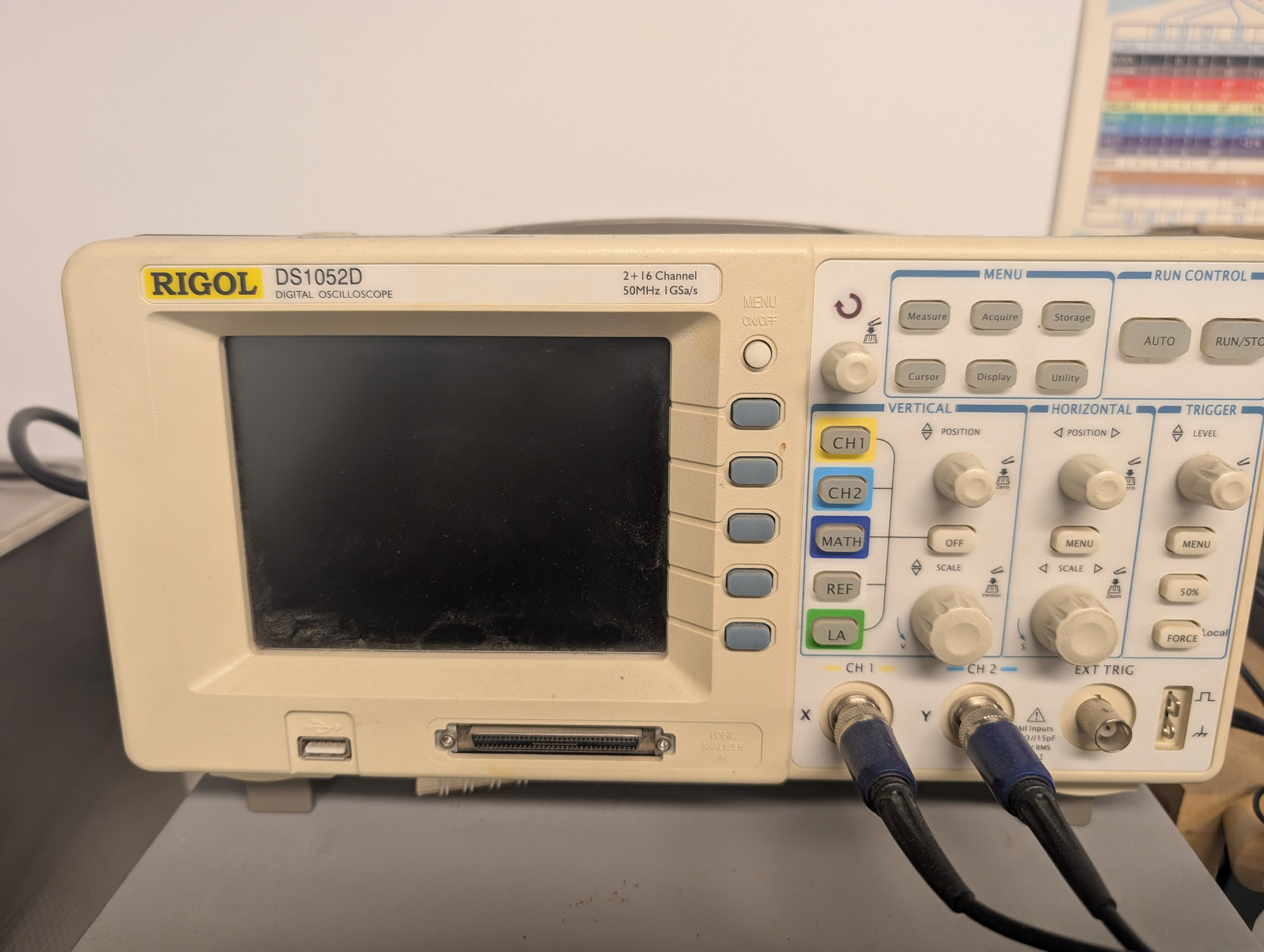 A beige LCD oscilloscope with "Rigol DS1052D 2+16 Channel 50MHz 1Gs/s" written at the top above the screen, and a USB port beside a SCSI-like connector under the screen. The right has two channel inputs, a trigger input, calibration connections, and an array of knobs and buttons.